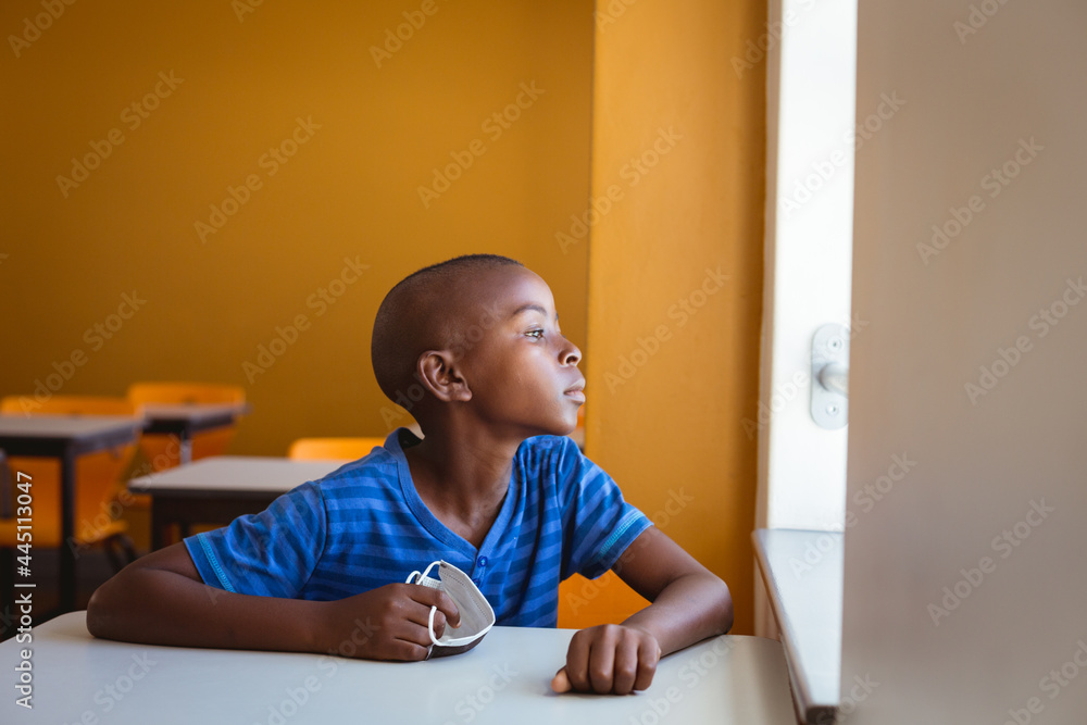 Fototapeta premium African american schoolboy holding face mask sitting at desk looking out of window in classroom