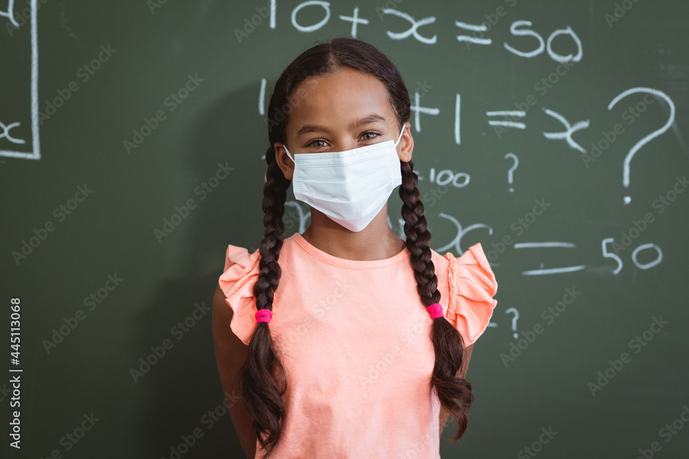 Fototapeta premium Portrait of mixed race schoolgirl in face mask standing in front of chalkboard in maths classroom