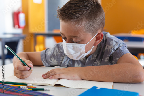 Caucasian schoolboy wearing face mask sitting at desk in classroom writing in book