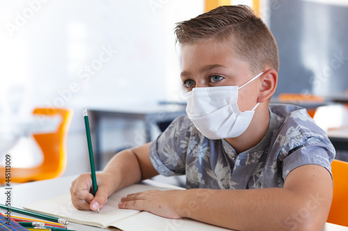 Caucasian schoolboy wearing face mask sitting at desk in classroom writing in book