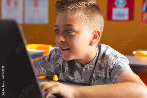Excited caucasian boy sitting at a desk in classroom using laptop and smiling