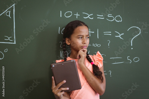 Mixed race schoolgirl standing in front of chalkboard in classroom using tablet and thinking