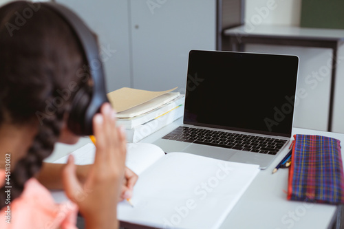 Mixed race schoolgirl in classroom wearing headphones and using laptop  with copy space on screen