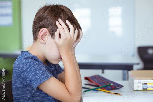 Tired caucasian schoolboy in classroom sitting at desk rubbing eyes