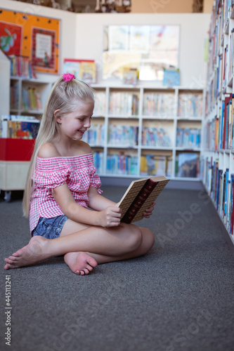 Smiling caucasian schoolgirl sitting on floor reading book in school library
