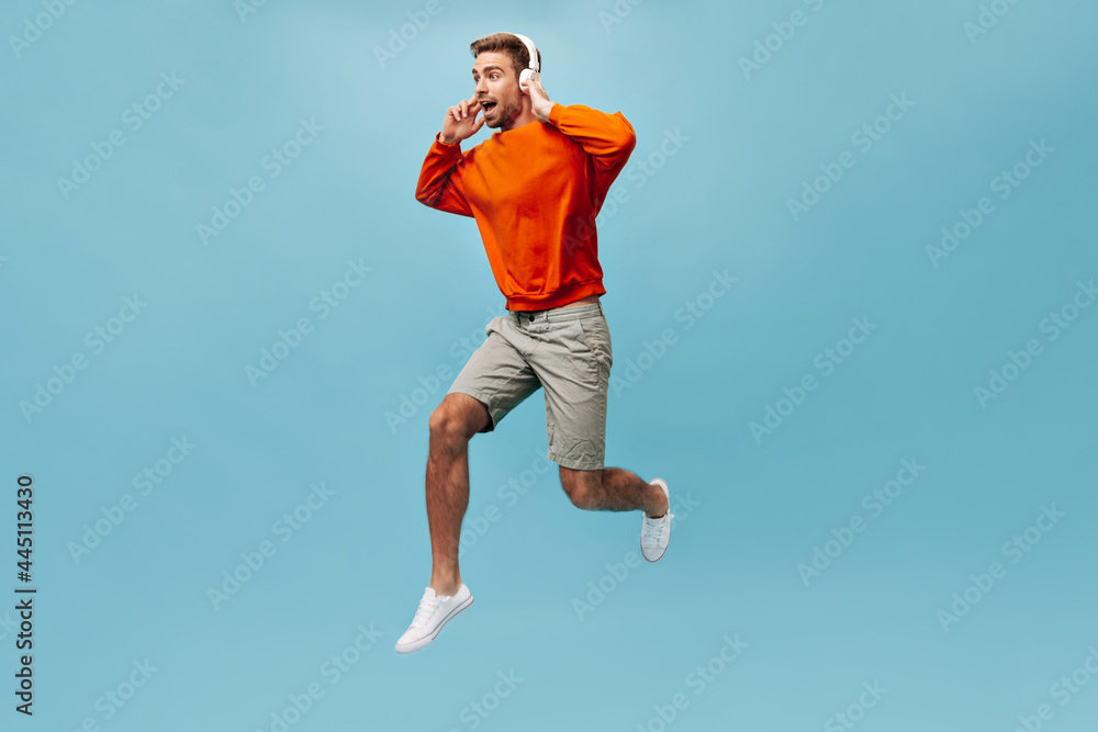 Optimistic brown haired man with beard in sweatshirt, shorts and sneakers holding white headphones and jumping on isolated background..