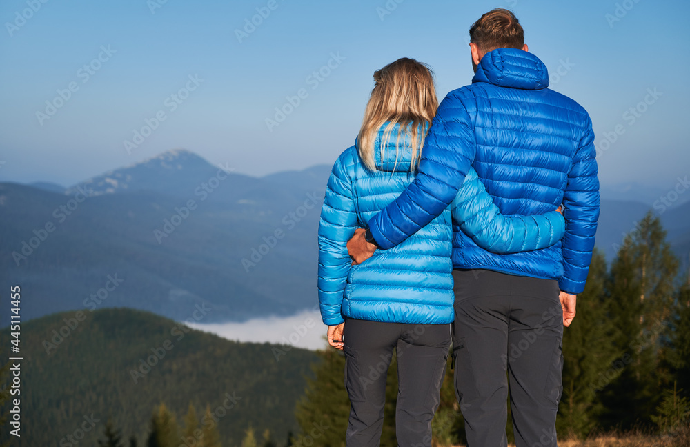 Rear view of couple hikers holding each other around waist and contemplating on fabulous mountain landscape in front of them at sunshine autumn vacation.