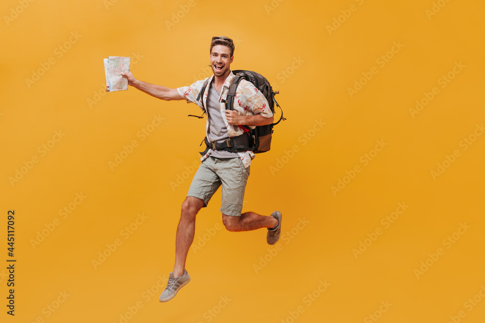 Slim happy man with beard in white summer shirt and grey shorts jumping with large backpack and map on isolated background.. Slim happy man with beard in white summer shirt and grey shorts jumping with large backpack and map on isolated background..