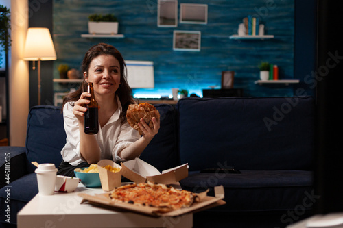 Happy woman holding beer bottle eating tasty takeaway food delivery while watching entertainment movie on television. Caucasian female enjoying fastfood home delivered sitting on sofa in evening