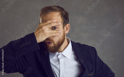 What a shame. Young business man or office worker in suit covering face with his hand and looking through fingers with one eye surprised and embarrassed by bad results or awkward situation at work photo