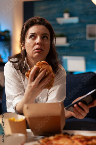 Fotografie Caucasian female holding tastu burgery in hands browsing on social media using smartphone relaxing on couch during junck-food night