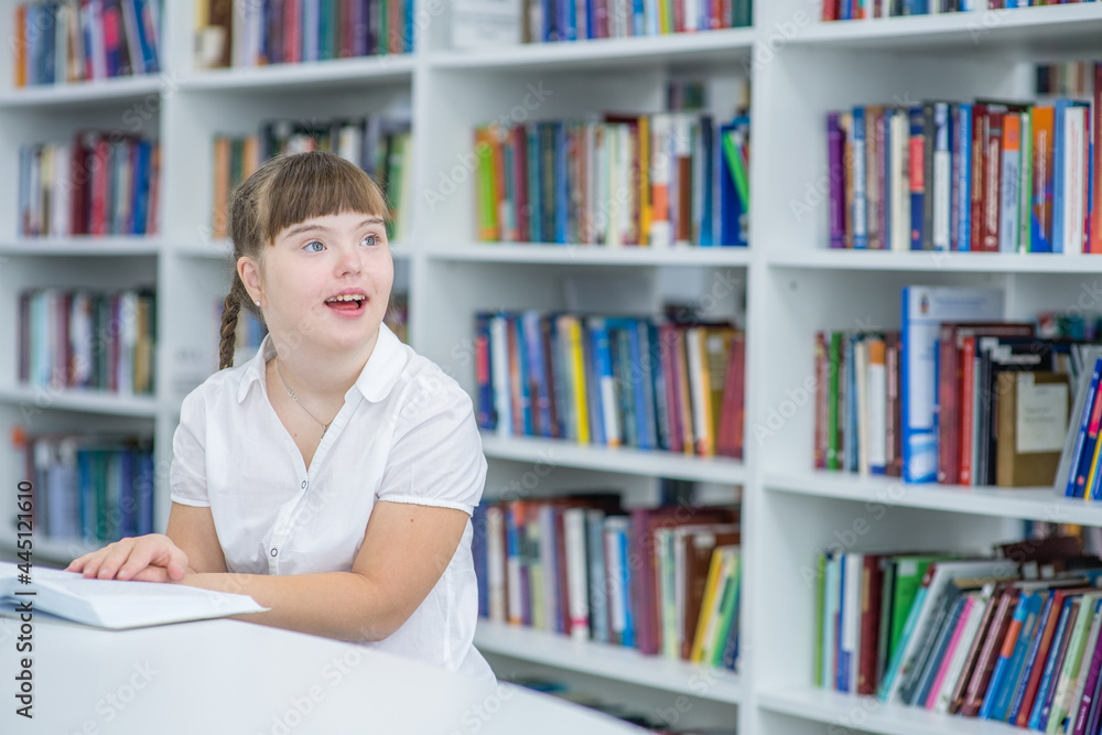 Happy young girl with syndrome down sits with a book at library. Education for disabled children concept. Empty space for text Happy young girl with syndrome down sits with a book at library. Education for disabled children concept. Empty space for text