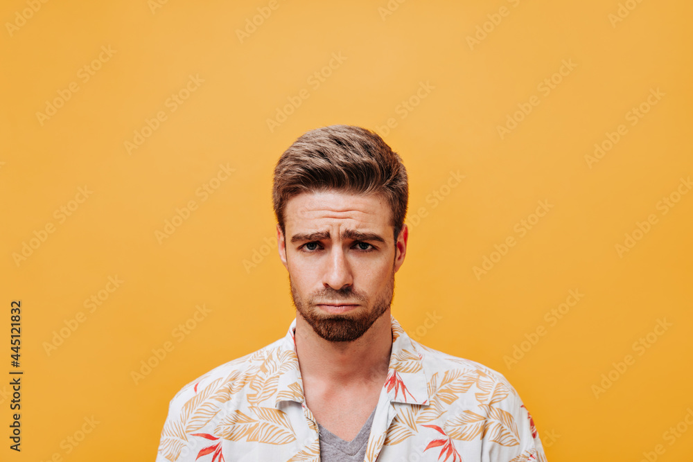 Disappointed man with stylish hairstyle and beard in trendy white outfit looking into camera on isolated orange backdrop.. Disappointed man with stylish hairstyle and beard in trendy white outfit looking into camera on isolated orange backdrop..