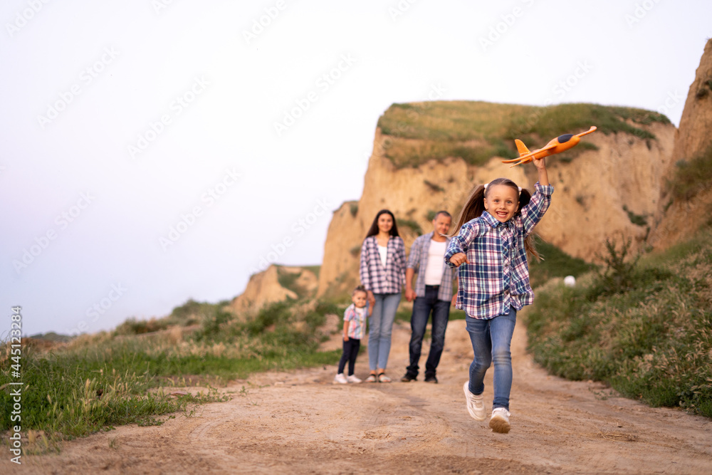 Foster family dad and mom with two daughters walking at sunset along a cliff or rocks in the summer evening. child launching airplane. happy parents and kids girls. Family adoption concept.