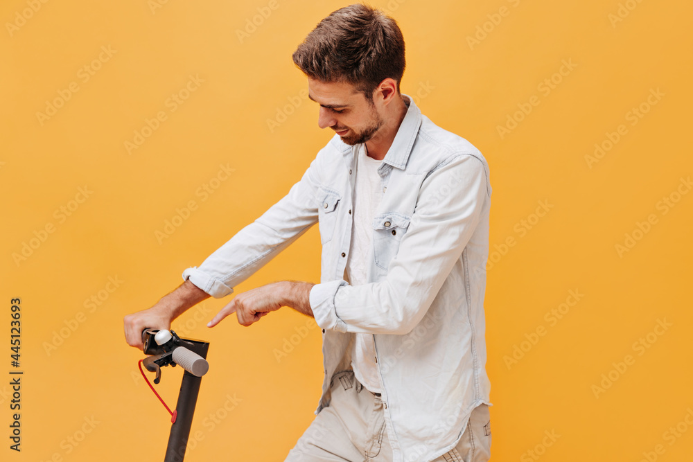Fototapeta premium Stylish bearded man in fashionable denim light clothes showing his finger on scooter and smiling on isolated orange background..