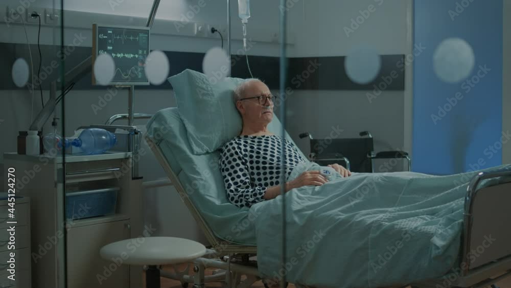 Ill patient with nasal oxygen tube and IV drip bag sitting in hospital ward bed to cure medical emergency, disease, recovery. Old man with oximeter ready for medicine, treatment Ill patient with nasal oxygen tube and IV drip bag sitting in hospital ward bed to cure medical emergency, disease, recovery. Old man with oximeter ready for medicine, treatment