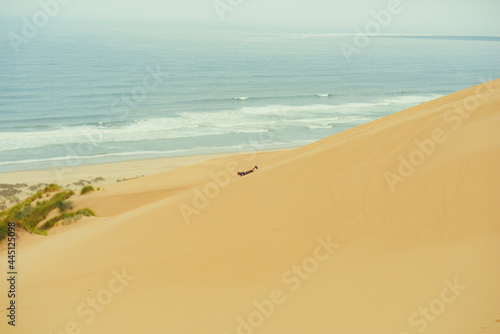 A man in shorts and a t-shirt sand boarding on a snowboard down the steep dune on his belly a wide view of the dunes and ocean in the background