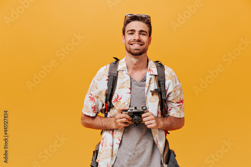 Smiling tourist with ginger beard in short sleeve summer shirt and plaid t-shirt holding camera and smiling on orange backdrop..