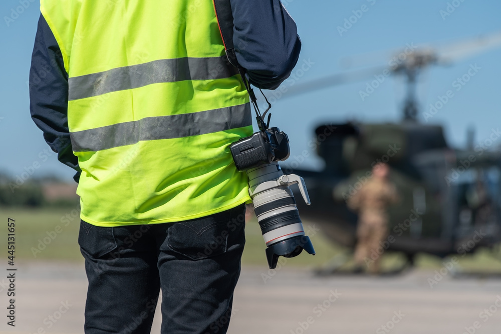 A military photographer, a reporter, in a signal yellow vest, ready to work in front of a military helicopter. Close-up. The background is blurred. A military photographer, a reporter, in a signal yellow vest, ready to work in front of a military helicopter. Close-up. The background is blurred.