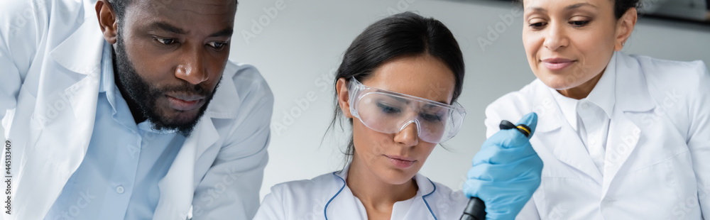 Scientist in goggles holding pipette near interracial colleagues, banner Scientist in goggles holding pipette near interracial colleagues, banner