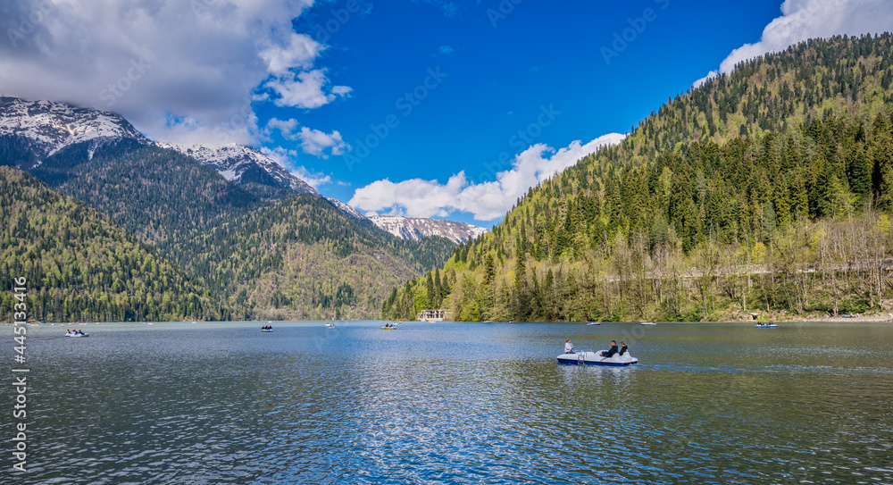 Beautiful view of Ritsa lake in Abkhazia with boats and catamarans against the snowy peaks of Caucasian mountains. Located at an altitude of 950 meters above sea level. Tourism, scenic destinations.