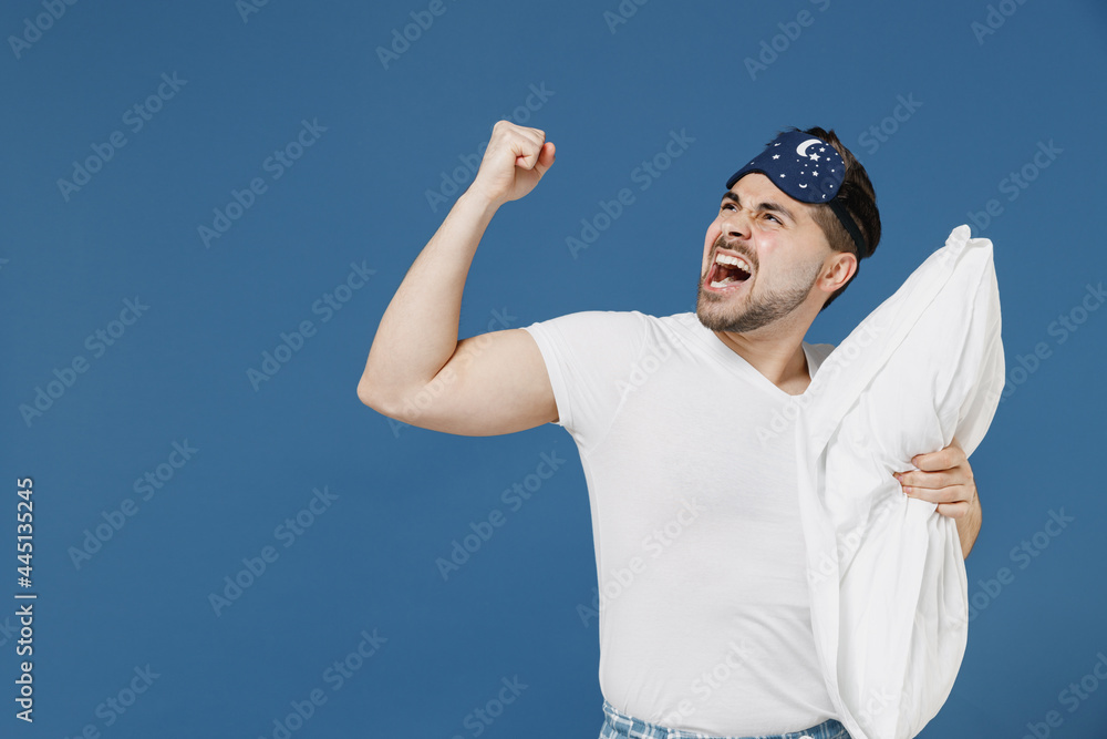 Naklejka premium Young stressed threatening annoyed sad tired man in pajama jam sleep mask rest relax at home hold pillow scream look aside clench fist isolated on dark blue background Bad mood night bedtime concept