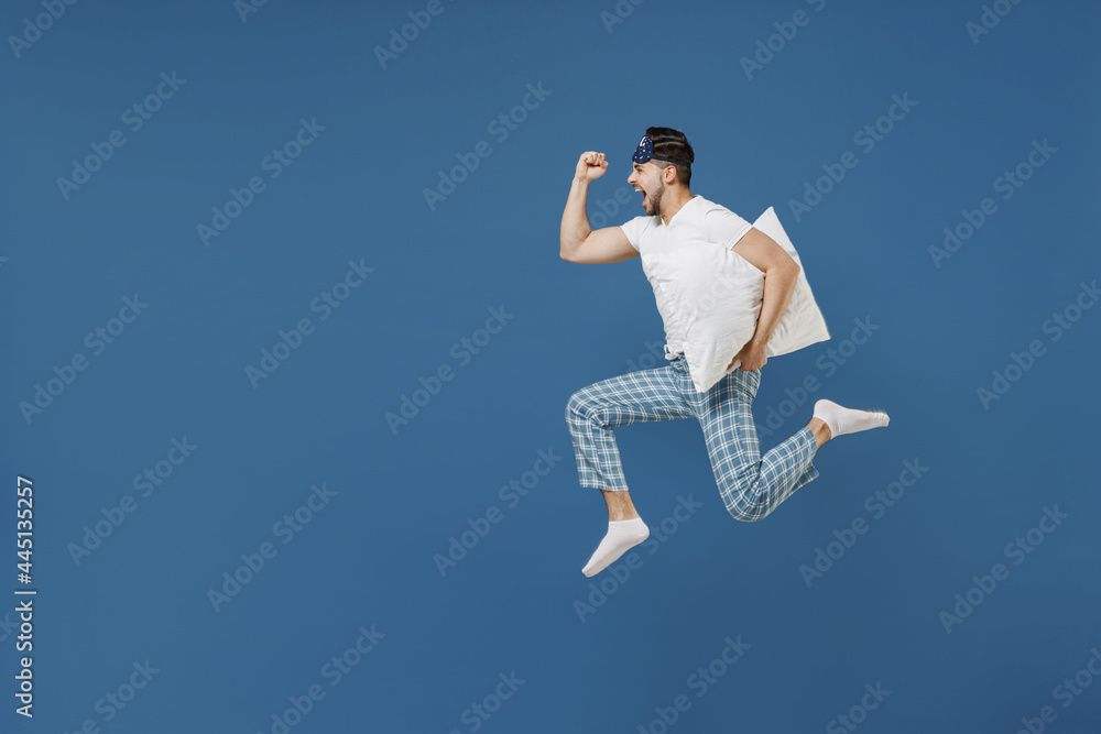 Full length side view young man 20s wearing pajamas jam sleep mask resting relaxing at home jump high hold pillow run fast hurry up isolated on dark blue background. Good mood night bedtime concept Full length side view young man 20s wearing pajamas jam sleep mask resting relaxing at home jump high hold pillow run fast hurry up isolated on dark blue background. Good mood night bedtime concept