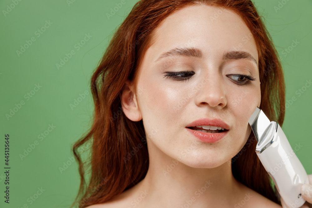 Fototapeta premium Beautiful close up half naked topless redhead hair woman 20s nude makeup clean face ultrasonic face scrubber isolated on pastel green color background Skin care healthcare cosmetic procedures concept