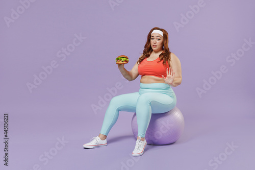 Full length fun young chubby overweight plus size big fat woman in red top warm up train sit on fit ball hold fast food burger cover with hand isolated on purple background gym Workout sport concept