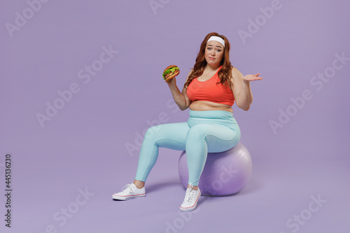Full length addicted young chubby overweight plus size big fat woman in top warm up train sit on fit ball biting fast food burger spread hand isolated on purple background gym Workout sport concept