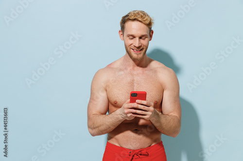 Handsome young sexy blond smiling fun man 20s in red shorts swimsuit relax near hotel pool hold using mobile cell phone isolated on pastel blue background. Summer vacation sea rest sun tan concept photo