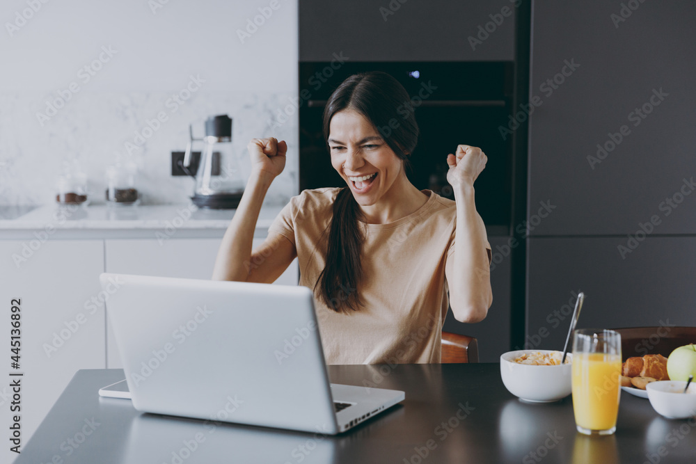 Young excited fun housewife woman 20s in beige shirt eat breakfast using laptop pc computer look for recipe do winner gesture cook food in light kitchen at home alone. Healthy diet lifestyle concept. Young excited fun housewife woman 20s in beige shirt eat breakfast using laptop pc computer look for recipe do winner gesture cook food in light kitchen at home alone. Healthy diet lifestyle concept.