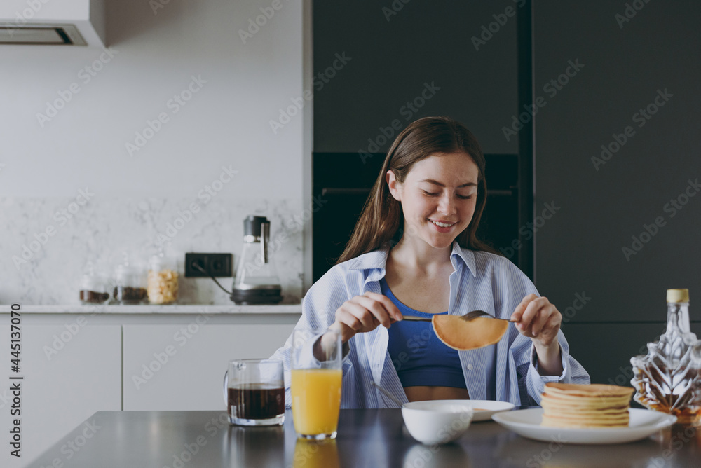 Young smiling fun satisfied happy housewife woman 20s wear casual clothes blue shirt eat breakfast pancakes with maple syrup cooking food in light kitchen at home alone Healthy diet lifestyle concept. Young smiling fun satisfied happy housewife woman 20s wear casual clothes blue shirt eat breakfast pancakes with maple syrup cooking food in light kitchen at home alone Healthy diet lifestyle concept.