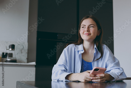 Minded pensive student young housewife woman 20s in blue shirt casual clothes drink using mobile cell phone look aside read recipe cook food in light kitchen at home. Healthy diet lifestyle concept photo