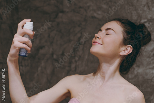 Foto Half naked topless young woman 20s apply on fixative and make-up base on face with closed eye doing morning routine in bathroom brown dark background