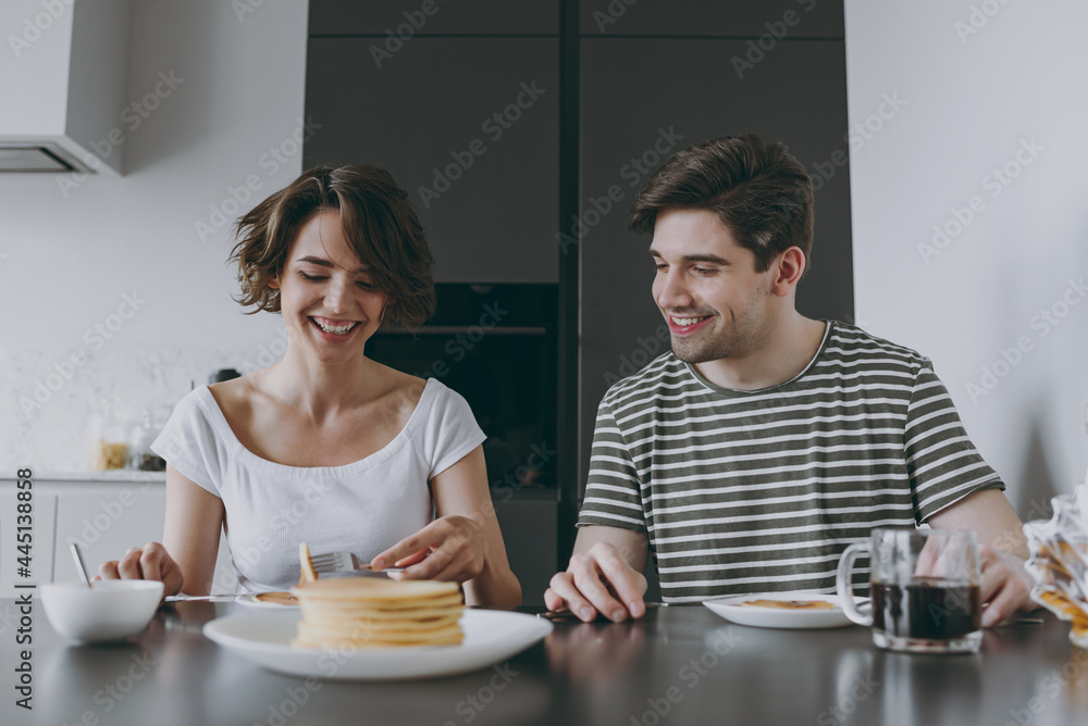 Young smiling fun happy couple two woman man in casual t-shirt clothes sit by table eat pancakes with maple syrup in morning cook food in light kitchen at home together Healthy diet lifestyle concept. Young smiling fun happy couple two woman man in casual t-shirt clothes sit by table eat pancakes with maple syrup in morning cook food in light kitchen at home together Healthy diet lifestyle concept.