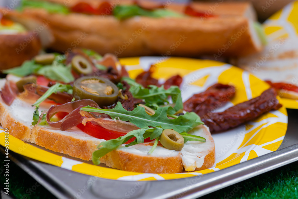 Sandwich with arugula, ham, cheese, jalapeno, sundried tomato on plate on counter at summer local food market - close up. Gastronomy, street food, mediterranean and Italian dish concept