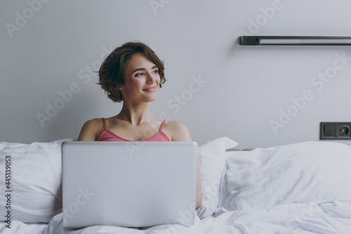 Calm pensive young woman in pajamas lies in bed wrap covered under blanket duvet on pillow hold use work on laprtop pc computer look aside rest relax indoors at home Good mood morning bedtime concept photo