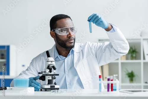 African american scientist in latex gloves and safety goggles holding test tube near microscope