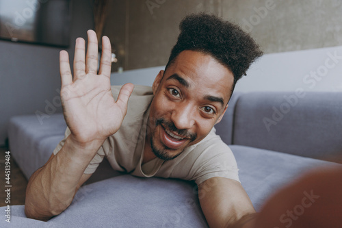 Close up full length young african american man in t-shirt sweatpants lay down on grey sofa waving hand talk by video call indoors partment do selfie shot on mobile phone rest on weekend stay at home photo