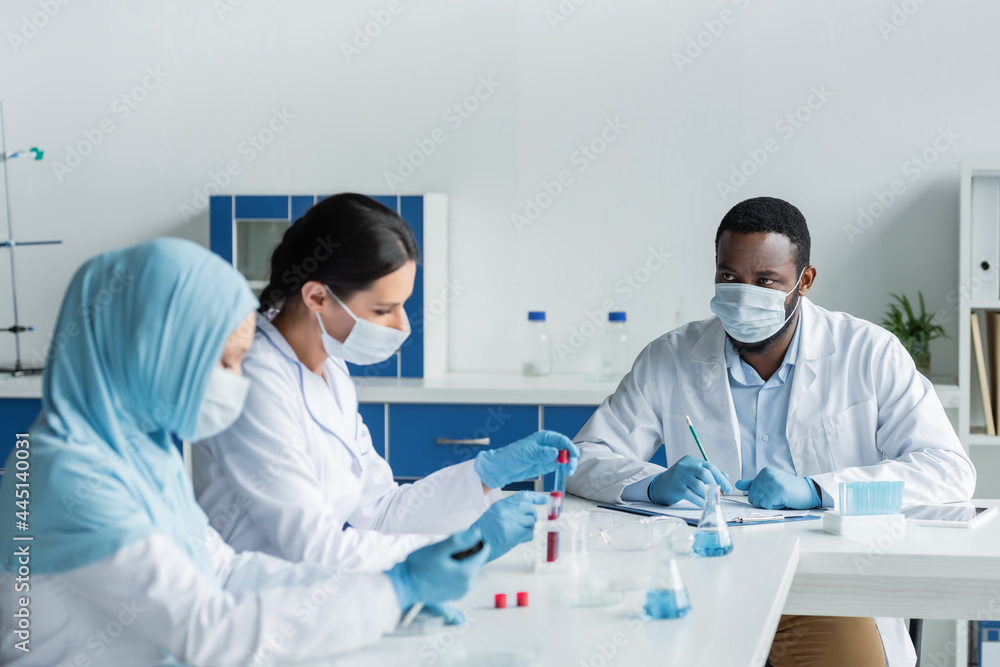 African american scientist with clipboard looking at blurred interracial colleagues with test tubes African american scientist with clipboard looking at blurred interracial colleagues with test tubes