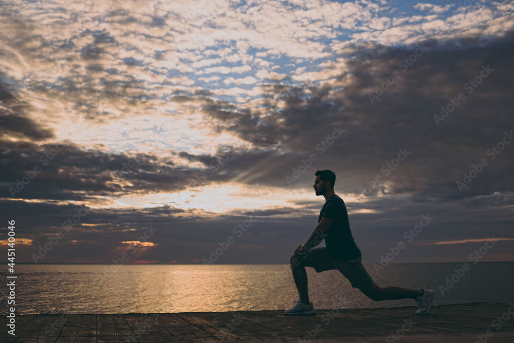 Full length side view young strong sporty athletic toned fit sportsman man in sport clothes warm up training do lunges at sunrise sun dawn over sea beach outdoor on pier seaside in summer day morning