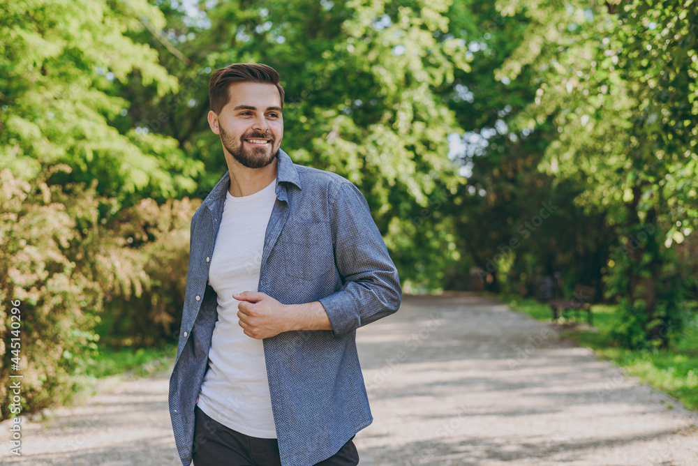 Young satisfied smiling happy man in blue casual shirt t-shirt walking look aside rest relax in spring green city park go down alley sunshine lawn outdoors on nature Urban lifestyle leisure concept. Young satisfied smiling happy man in blue casual shirt t-shirt walking look aside rest relax in spring green city park go down alley sunshine lawn outdoors on nature Urban lifestyle leisure concept.