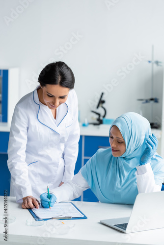 Smiling arabian scientist writing on clipboard near laptop, colleague and petri dishes