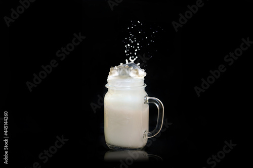 Milk splashes out of a transparent square jar of a mug  flies in all directions  falls on the table on a black background.