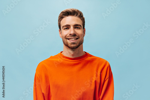Positive young guy with red beard in bright orange sweatshirt looking into camera and smiling on isolated blue background.. photo