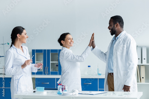 Smiling african american scientists giving high five near colleague with safety goggles in lab