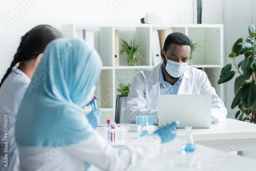 African american scientist in medical mask using laptop near blurred colleagues with equipment