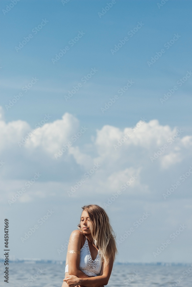 A young tanned slim beautiful blonde woman in a white knitted top, with a gold boho sticker on arm on the background of the sea, sky with clouds. Hot sunny day. Music album cover, free space for text