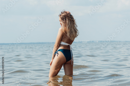 A young tanned slim beautiful blonde woman in a swimwear poses on the background of the sea and sky. A sexy girl is standing with her back lifting hair. The light from the water is reflected on body